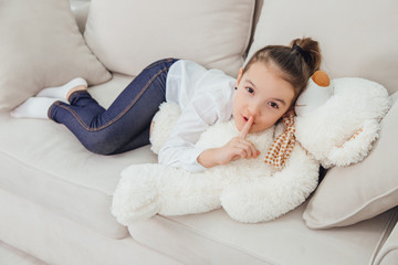 Nice little girl is looking at the camera, showing a silence sign while lying on her big white teddy-bear. Full-length.