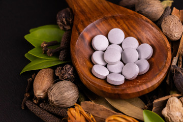 High angle closeup shot of white herbal pills in a wooden serving spoon with green and dry herbs on black background. Herbal medicine concept