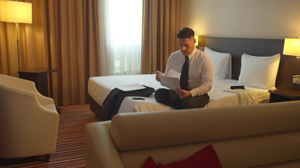 Man in shirt and tie sit on the bed in hotel and write on paper
