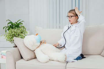 Pretty little girl wearing doctor coat, sitting on the sofa with legs curled up, examining her sick teddy-bear with stethoscope.