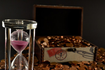 Hourglass full of pink sand and chest full of coins in a background. Isolated on a black background