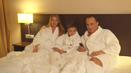 Obraz premium Parents and little son sit on bed and watch TV in hotel room