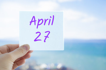 April 27th. Hand holding sticker with text April 27 on the blurred background of the sea and sky. Copy space for text. Spring month in calendar concept