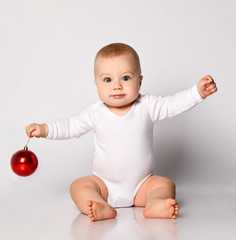 Cheerful infant baby boy toddler in white bodysuit sitting holding handed playing with red...