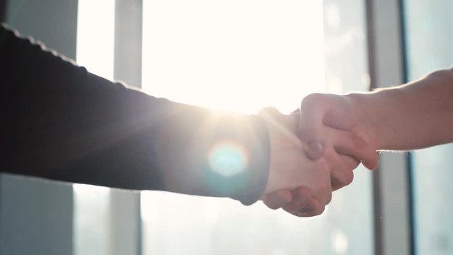Close-up of two businessmen shaking hands in the office against the background of a window and sunset. Camera makes sharp turn, emphasizing importance of the moment. Tracking shot in slow motion.
