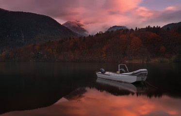 Fototapeta na wymiar Autumn Landscape With Two White Boats Of The Rescue Service Against The Backdrop Of The Reflection Of Triglav Mountains, Fiery Red Beech Forest And Enchanting Pink Dawn Sky. Lake Bohinj, Slovenia.