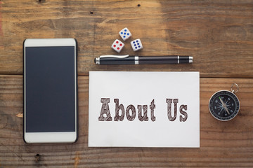 About Us Words written on white on wooden desk with dice,compass,smart phone and pen.Top view conceptual