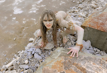 A young woman is  dressed as a neanderthal warrior.  She is covered with mud, filth and dirt and is seen in  a stone quarry area surrounding.
