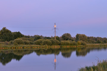 The power transmission tower and the moon are visible on the lake in the evening. 