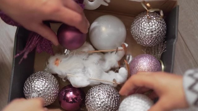 Close-up scene of a box with a christmas toys. There are white, silver and purple balls. Four hands are taking four balls to hang on a christmas tree.