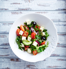 Healthy salad with black olives, feta cheese, cherry tomatoes, cucumber and rocket on bright wooden background. Top view. 