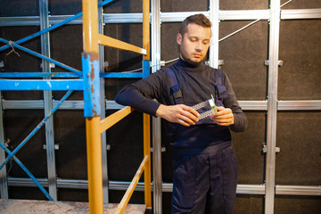 Worker stands near the wall and hold metal detail in hand. Worker pondering the next step of construction process.