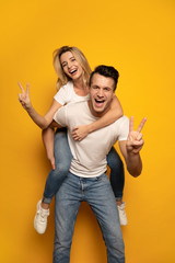 Peace and love! Full-length photo of a cheerful man, who is giving his wife a piggyback ride, both are laughing with joy and showing peace signs to the camera.