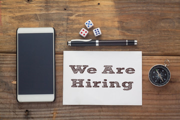 We Are Hiring Words written on white on wooden desk with dice,compass,smart phone and pen.Top view conceptual