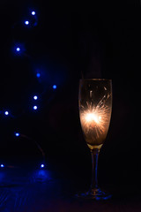 Sparkler inside a glass of champagne, which stands on a wooden table in the dark, in the background are blue bokeh lights.