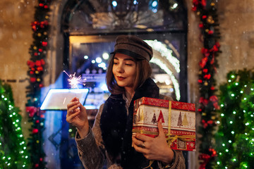 Christmas, New Year concept. Woman burning sparkler on city street by decorations. Girl holding gift box under snow.