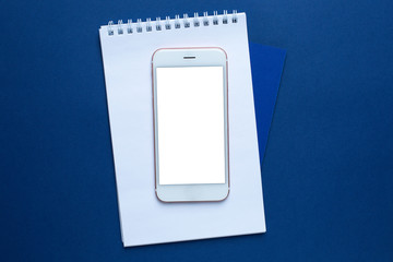 Mobile phone and white office notebook on a classic blue background with copyspace. Trendy Pantone color 2020