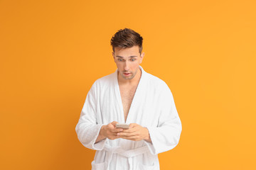 Shocked man in bathrobe and with mobile phone on color background