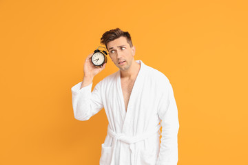 Emotional man in bathrobe and with alarm clock on color background