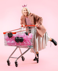 Cool fashionable mature woman with air balloons in shopping cart on color background