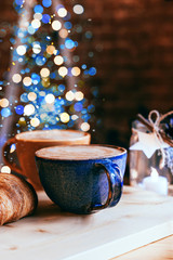 Perfect breakfast of croissants and cappuccino in a cafe. Blue and yellow ceramic cups of cappuccino and croissants on wooden table with Christmas lights and bokeh background. Close-up.