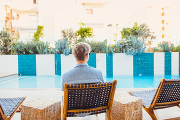 Back view young caucasian man in blue shirt relaxing on chair near swimming pool watching running water and plant flowebed. Summer, travel, vacation and holiday concept. Slow lifestyle. Copy space.