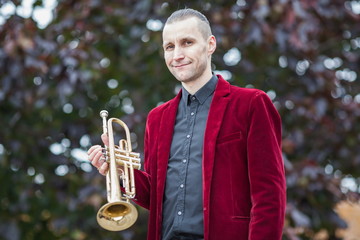 young attractive musician in red velvet jacket and black shirt smiles and holds golden trumpet or...