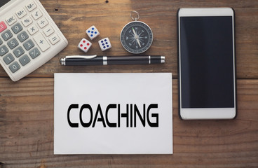 Coaching  written on paper,Wooden background desk with calculator,dice,compass,smart phone and pen.Top view conceptual.