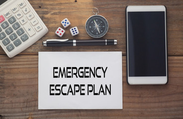 Emergency Escape Plan  written on paper,Wooden background desk with calculator,dice,compass,smart phone and pen.Top view conceptual.