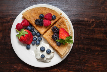 Pancakes with berries and sour cream on a dark background. Food background. Healthy and delicious breakfast. Sweet pancakes in a white plate on a wooden table. Top view