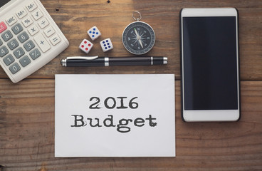 2016 Budget written on paper,Wooden background desk with calculator,dice,compass,smart phone and pen.Top view conceptual.