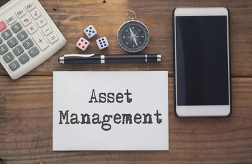 Asset Management written on paper,Wooden background desk with calculator,dice,compass,smart phone and pen.Top view conceptual.