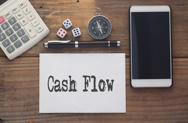 Cash Flow written on paper,Wooden background desk with calculator,dice,compass,smart phone and pen.Top view conceptual.