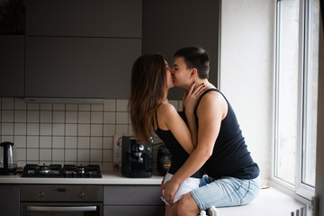 Young couple of millennials in black t-shirts, jeans hugging in the kitchen in the morning by the window. Loving lovers have fun before breakfast at home. Man sits on windowsill, kisses woman standing