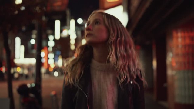 Young blonde woman in jeans and leather jacket posing and walking on streets in downtown after sunset in the evening