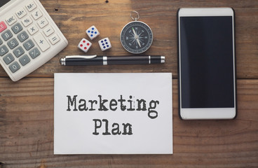 Marketing Plan written on paper,Wooden background desk with calculator,dice,compass,smart phone and pen.Top view conceptual.