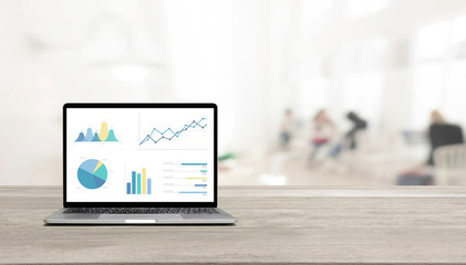 Modern laptop on wooden table showing charts and graph against office with white wall and windows...