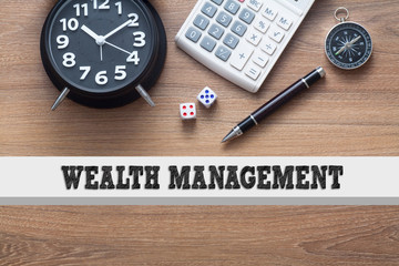 WEALTH MANAGEMENT written conceptual,alarm clock,dice,pen,calculator and compass on wood background