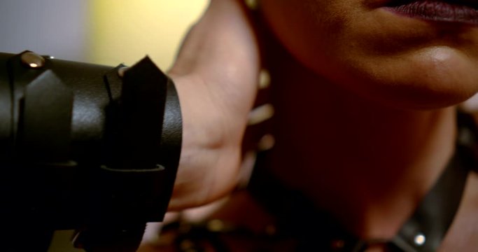 a hand in leather straps touches the neck of a blonde with a bright make-up and a gunbelt. close up. contoured pink light. dark key