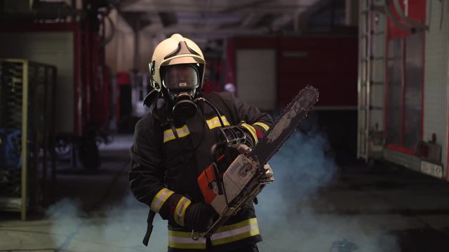 Firefighter portrait wearing full equipment, oxygen mask, and chain saw. Smoke and fire trucks in the background.