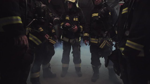 group of five firefighters wearing full equipment. relaxed chatting after work well done. smoke and fire trucks in the background. Name tag translation is fireman and model's last name.
