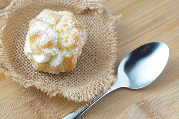A close up of a creampuff with an ising on top. Placed on a piece of sack next to a silver spoon  over a wooden background