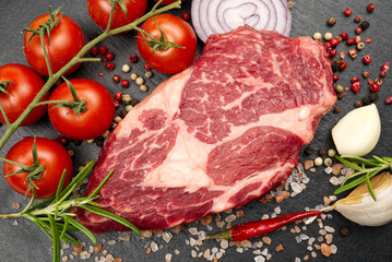 Raw rib eye steak with spices and vegetables. Ingredients for restaurant meal. Fresh meat, salt, rosemary, thyme, chilli, cherry tomatoes, garlic on black stone. Food background.