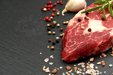 Raw rib eye steak with spices and vegetables. Ingredients for restaurant meal. Fresh meat, salt, rosemary, thyme, chilli, cherry tomatoes, garlic on black stone. Food background.