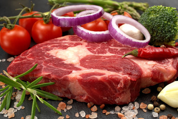 Raw rib eye steak with spices and vegetables. Ingredients for restaurant meal. Fresh meat, salt, rosemary, thyme, chilli, cherry tomatoes, garlic on black stone. Food background.