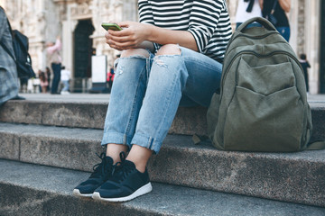 A girl in casual clothes a tourist or a student with a backpack uses a cell phone while sitting on the steps. Youth lifestyle and modern technology