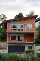 Narrow red building blocks suburban family house under construction with new windows and roof on side of small hill surrounded with garden vegetation and other houses on warm sunny summer day