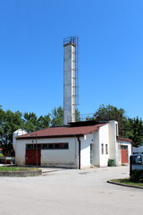 Small industrial building in front of tall narrow white rectangle concrete chimney with metal stairs secured with round steel safety tunnel mounted on one side surrounded with paved parking lot