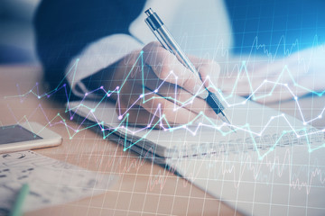 A woman hands writing information about stock market in notepad. Forex chart holograms in front....