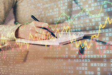 Hands writing information about stock market in notepad. Forex chart holograms in front. Concept of...